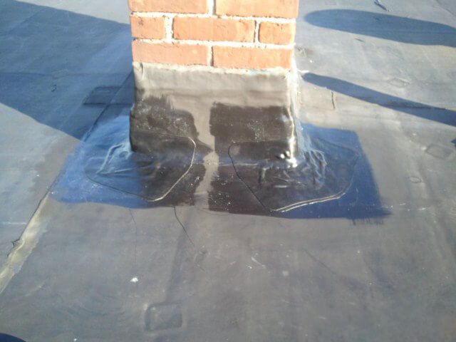 Black EPDM rubber roof with fresh flashing and sealant applied around the base of a brick chimney