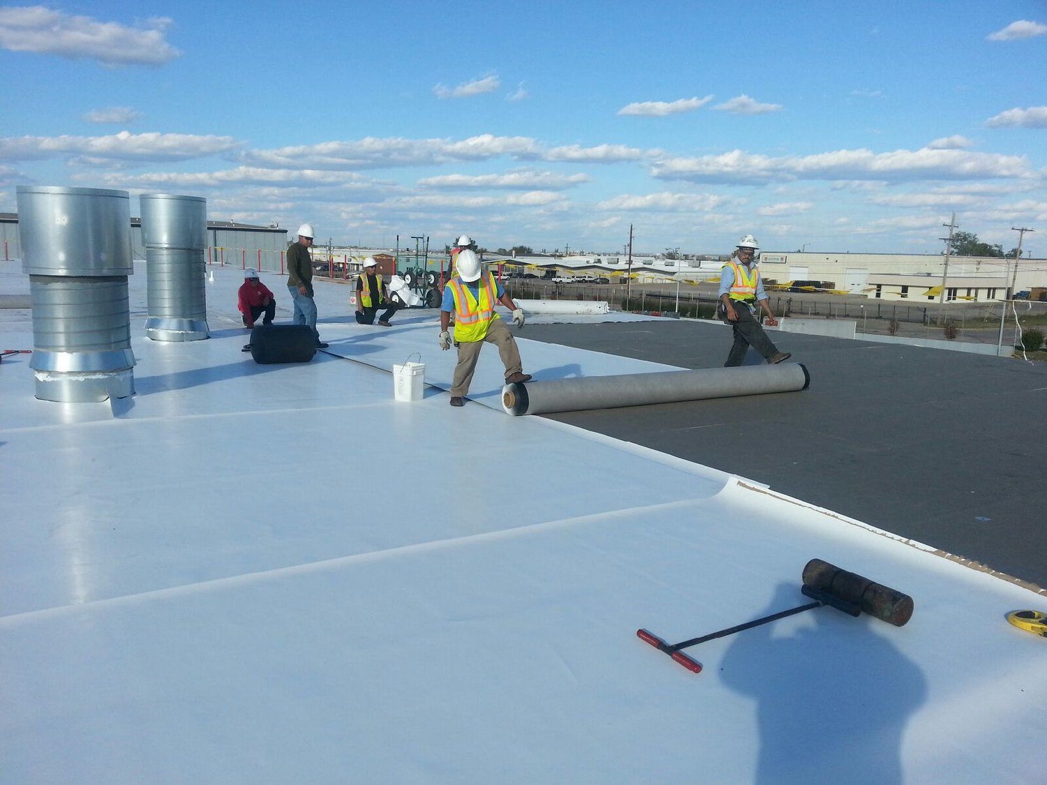 Roofing crew in hard hats and safety vests unrolling white TPO membrane across a large commercial flat roof