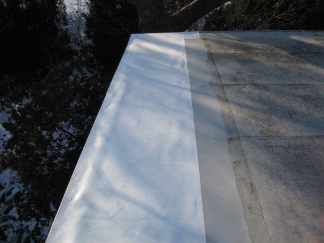 Edge of a white PVC flat roof in winter with snow on the ground below, showing a repaired section next to weathered membrane